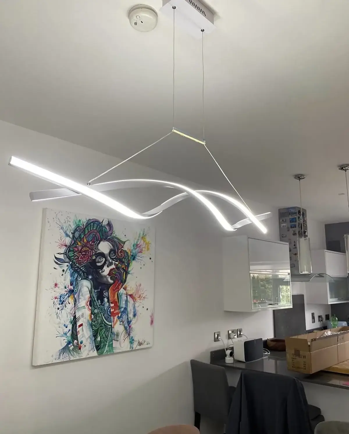 Two curved linear LED elements suspended on fine cables from a white ceiling above a kitchen counter, with a colourful painting on the wall