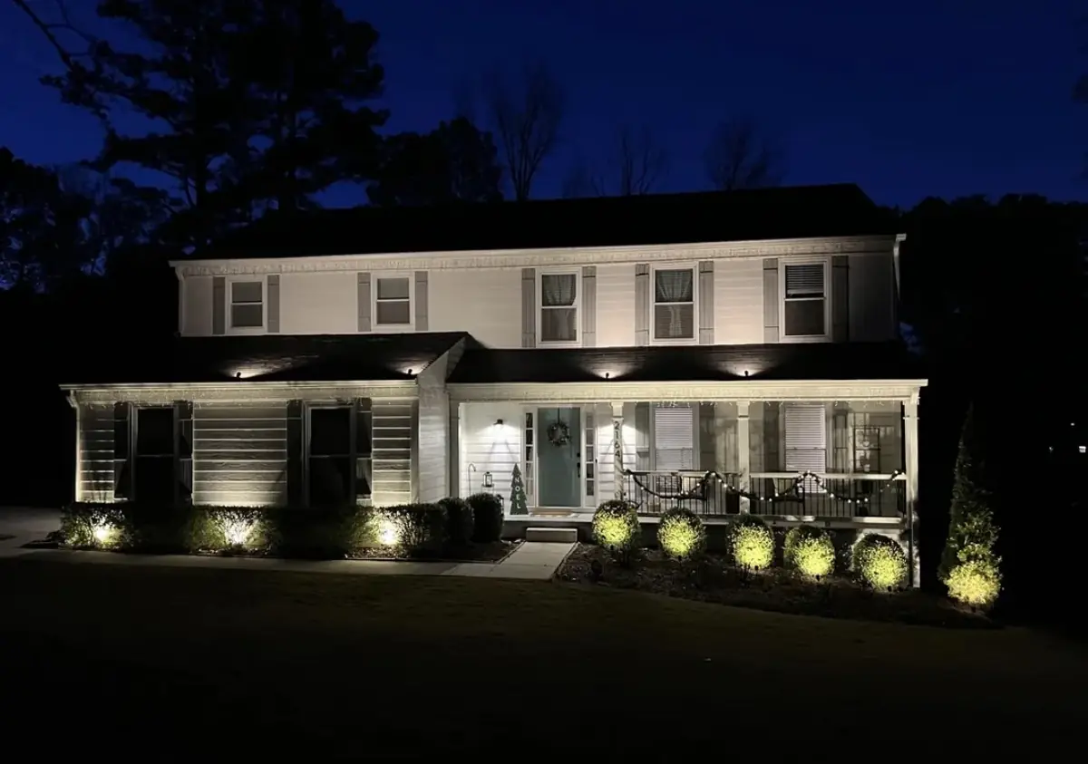 Two-storey white house at dusk under a deep blue sky, with downlights under the porch roof and uplights on the shrubs along the front